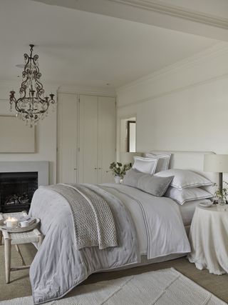white and gray bedroom with vintage chandelier, original features, wood stool, white rug, coir carpet, white and grey bedding, white bed