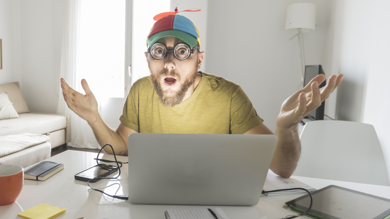 Bizarre man looking confused in front of a laptop at home with a colorful propeller hat on.