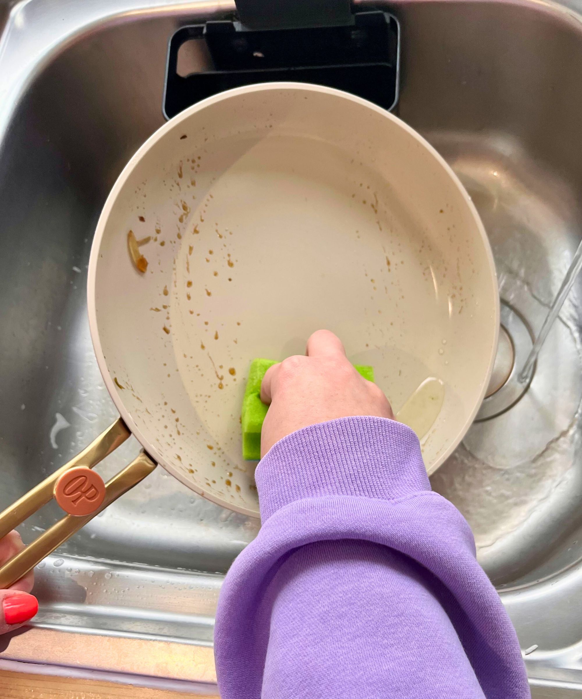 It was easy to hand-wash one of the pans in the Our Place 11-Piece Essentials Cookware Set, pictured here with cooking oils and stains, with a hand pulling a soft sponge down the middle and leaving no streaks or marks behind
