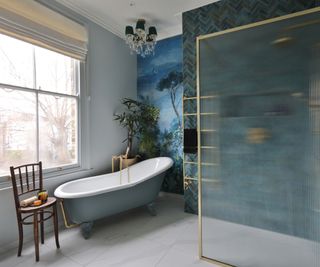 Freestanding roll-top bath beside a ribbed glass shower screen, with blue tiles, brass fittings and a mural-style feature wall.