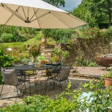 patio area in a garden with a large parasol, table and chairs, firepit and lots of planting and raised beds