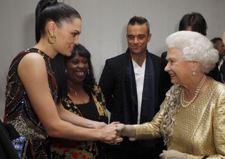 Queen Elizabeth wearing a gold dress shaking hands with Jessie J and smiling while Robbie Williams watches in the back