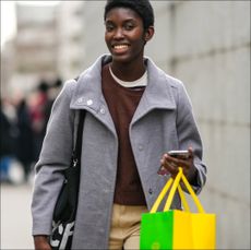 A model wears a white and navy blue striped print pattern t-shirt, a dark brown sweater, a pale gray long buttoned coat, beige large suit pants, a yellow and green shopping bag, outside Chloe, during Paris Fashion Week - Womenswear F/W 2022-2023, on March 03, 2022 in Paris, France