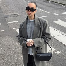 hetty wears gray wool coat, gray sweater, and black pants, and a black handbag while holding a to-to coffee cup near a crosswalk. 