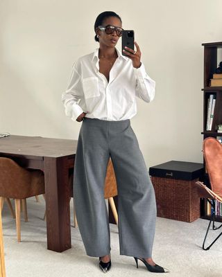 @nlmarilyn taking a mirror selfie wearing a white button-down, baggy grey trousers, and black heels