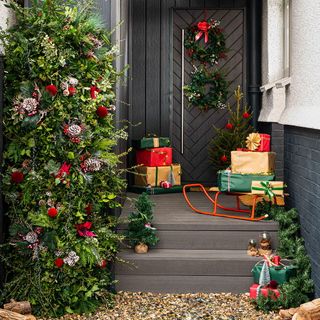 a black porch decorated with a large christmas wreath tower, two christmas wreaths on the door, a selection on red, gold and green wrapped presents on the porch and orange sleigh