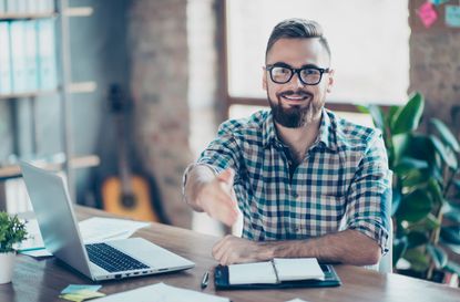 Concept of successful job interview. Portrait of happy cheerful smiling glad worker giving you a hand for handshaking, he is sitting at his comfortable office in front of laptop