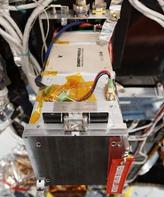 a small box wrapped in metallic foil on the side of a tall rectangular spacecraft on four legs