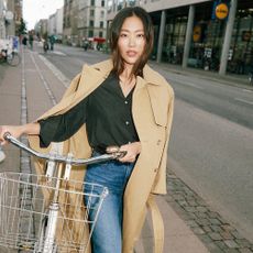A woman in a blakc shirt, jeans and a beige coat with a bike.
