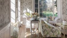 A floral living room with floral wallpaper, floral couch, white coffee table, rattan rug, wooden flooring, large glass vase with white flowers, and large windows out onto a leafy garden.