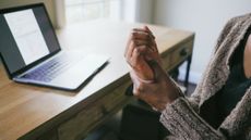 Woman rubs her palm with her thumb. She is sitting at a desk with an open laptop on it