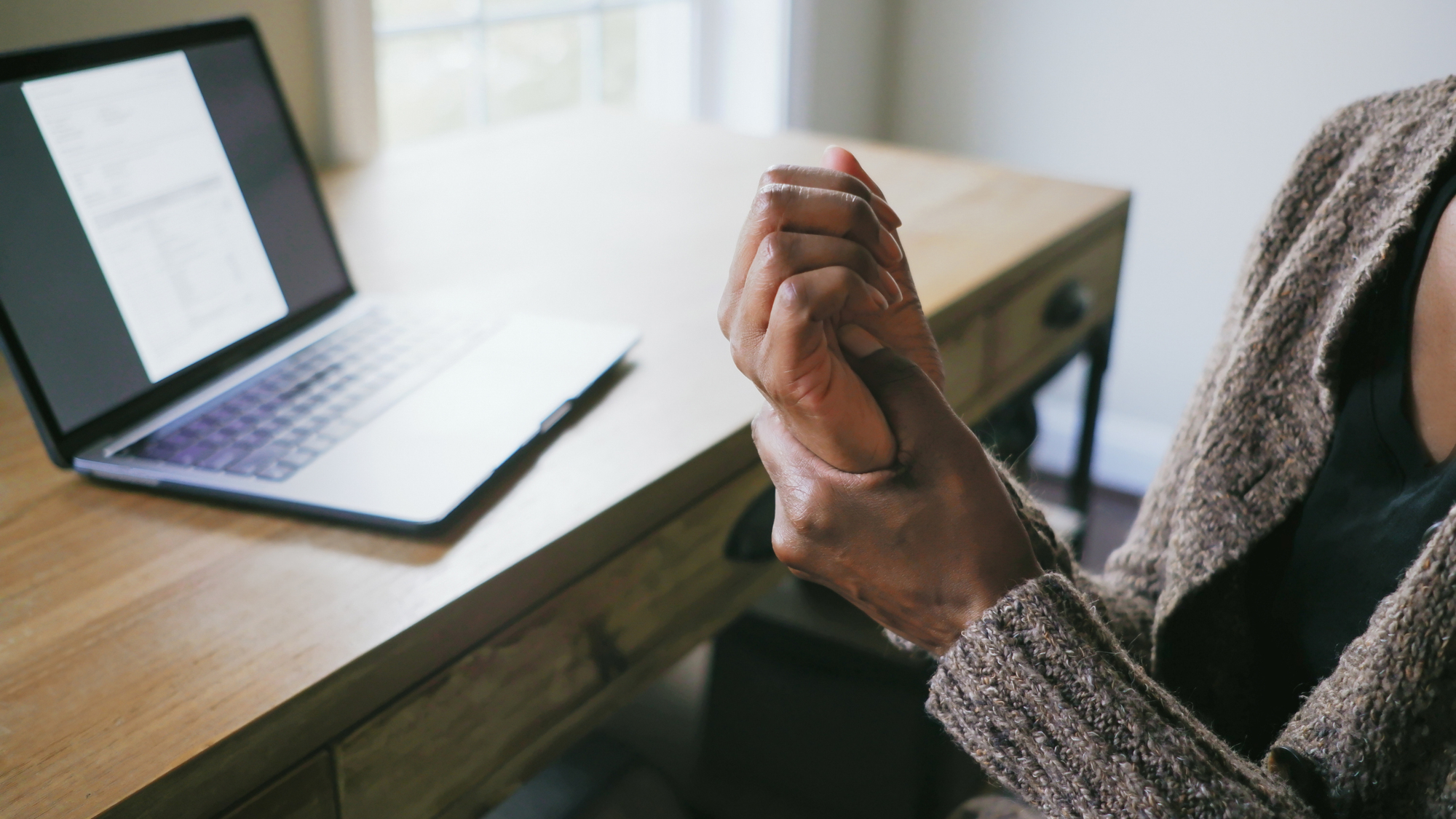 Woman rubs her palm with her thumb. She is sitting at a desk with an open laptop on it