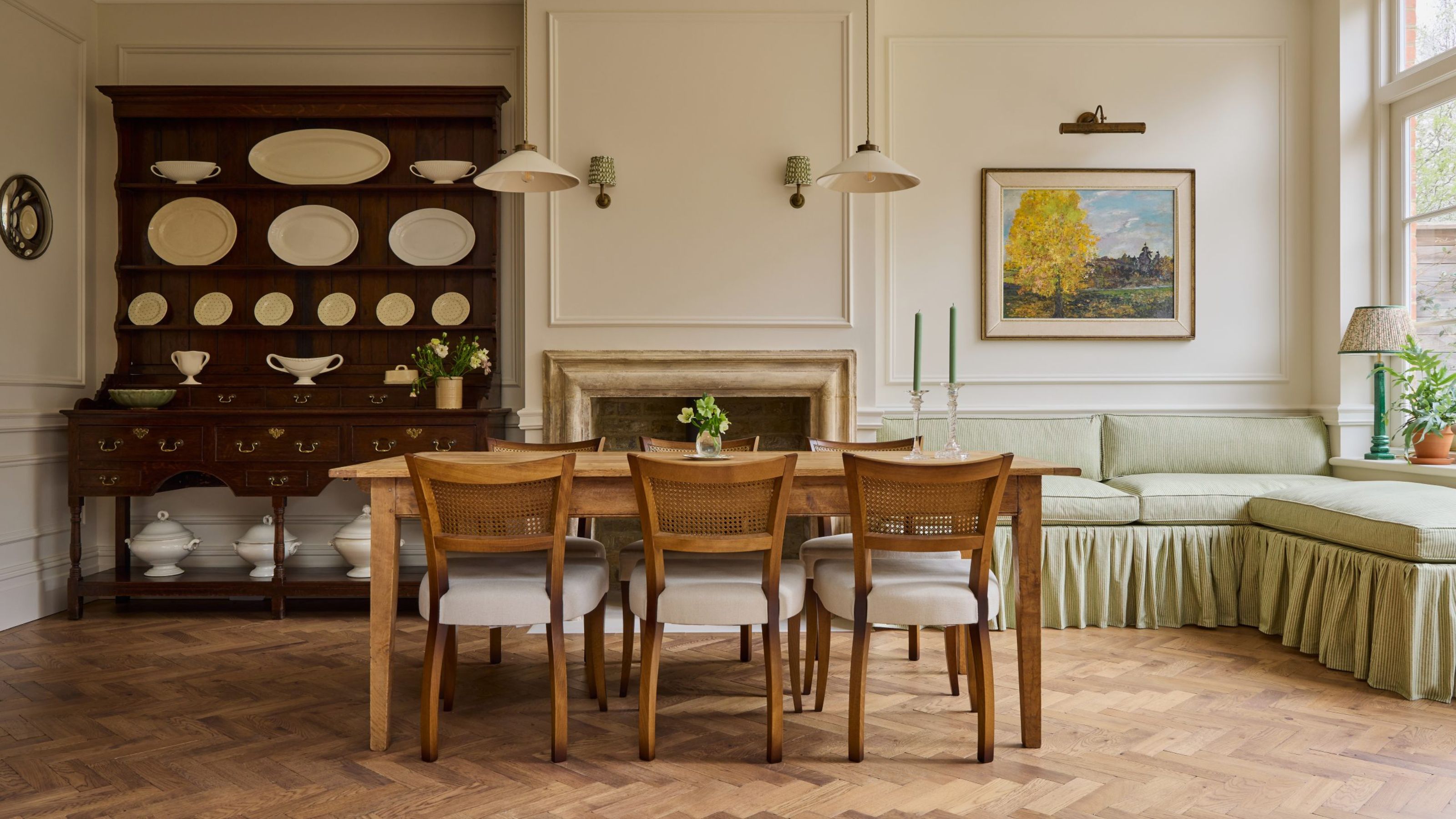 Elegant dining room with a wooden dining table, cane-back chairs, a dark antique hutch with white pottery, a stone fireplace, and a sage green corner bench on parquet floors.