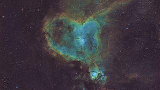A swirling deep space nebula is pictured that takes the vague shape of a cartoonish human heart. The centre of the heart nebula is a pale blue/teal, while the outside is formed of dense dusty filaments of yellow and brown. A colorful starfield is visible in the background.