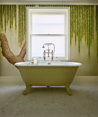 Large green bathroom with freestanding bath and large window to the back. There is a weeping willow tree wallpaper across the back wall