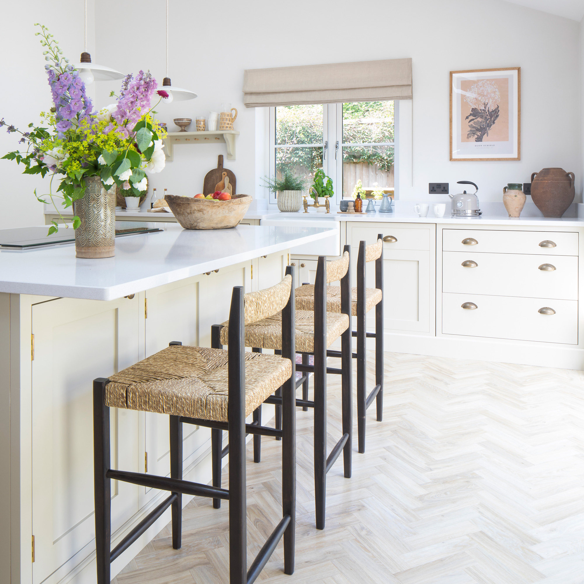 country cottage kitchen with cream coloured shaker cabinets and cup handles, a kitchen island with a vase of flowers and wicker bar stools upon timber herringbone flooring