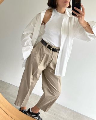 Die britische Mode-Influencerin Lucy Alston posiert für ein Selfie im Spiegel und trägt ein weißes Hemd mit Knopfleiste, ein weiß gestreiftes Tanktop, einen schwarzen Gürtel, kurze plissierte Khakis und schwarze Adidas Samba-Turnschuhe.