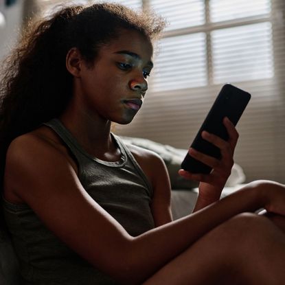 Side view of desperate teenager with cellphone sitting by bed in her room - stock photo