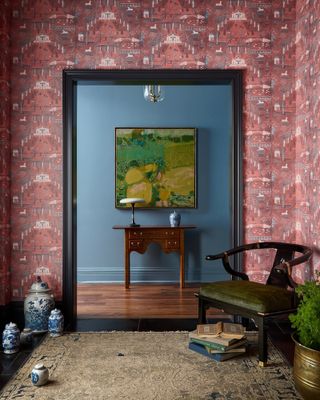 View from living room with muted red patterned wallpaper into entryway with mid-century blue walls, dark wood console and framed green and yellow artwork