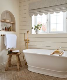 A neutral bathroom with terracotta floor tiles, a white freestanding bathtub, and scallop edge blinds