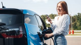 woman traveling by electric car having stop at charging station standing plugging cable browsing internet on smartphone