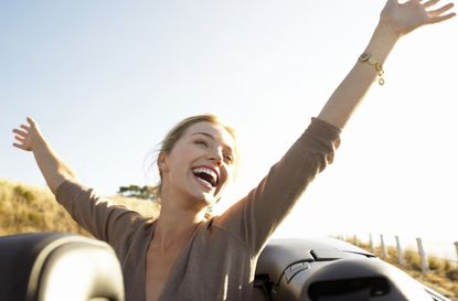 Young Woman Sits in the Back of a Convertible, Her Arms in the Air, Laughing With Joy