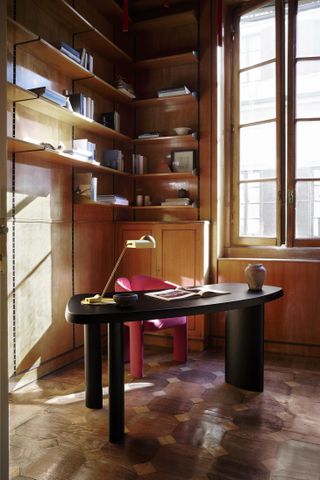 A black wooden desk by Charlotte Perriand, featuring a curved top and photographed in a studio by a window and near a series of wooden bookshelves