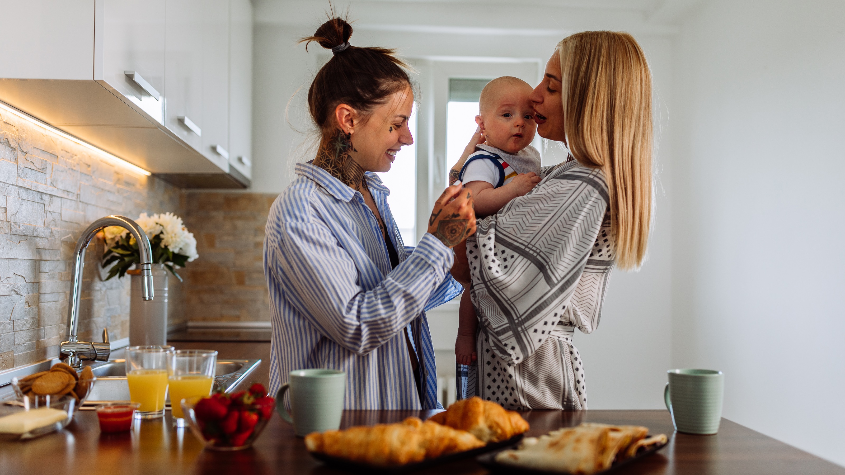 Two young women cuddle with their baby boy in their kitchen.