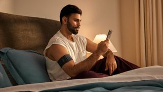 A man in white vest sitting up in bed checking phone with Garmin Index Sleep Monitor on right arm