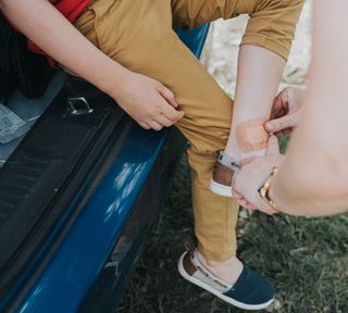 an adult putting a band aid on boy's ankle at car - GettyImages-1240331062