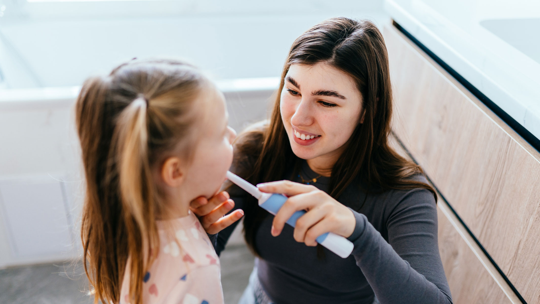 A picture of a woman helping a young girl brush her teeth with an electric toothbrush