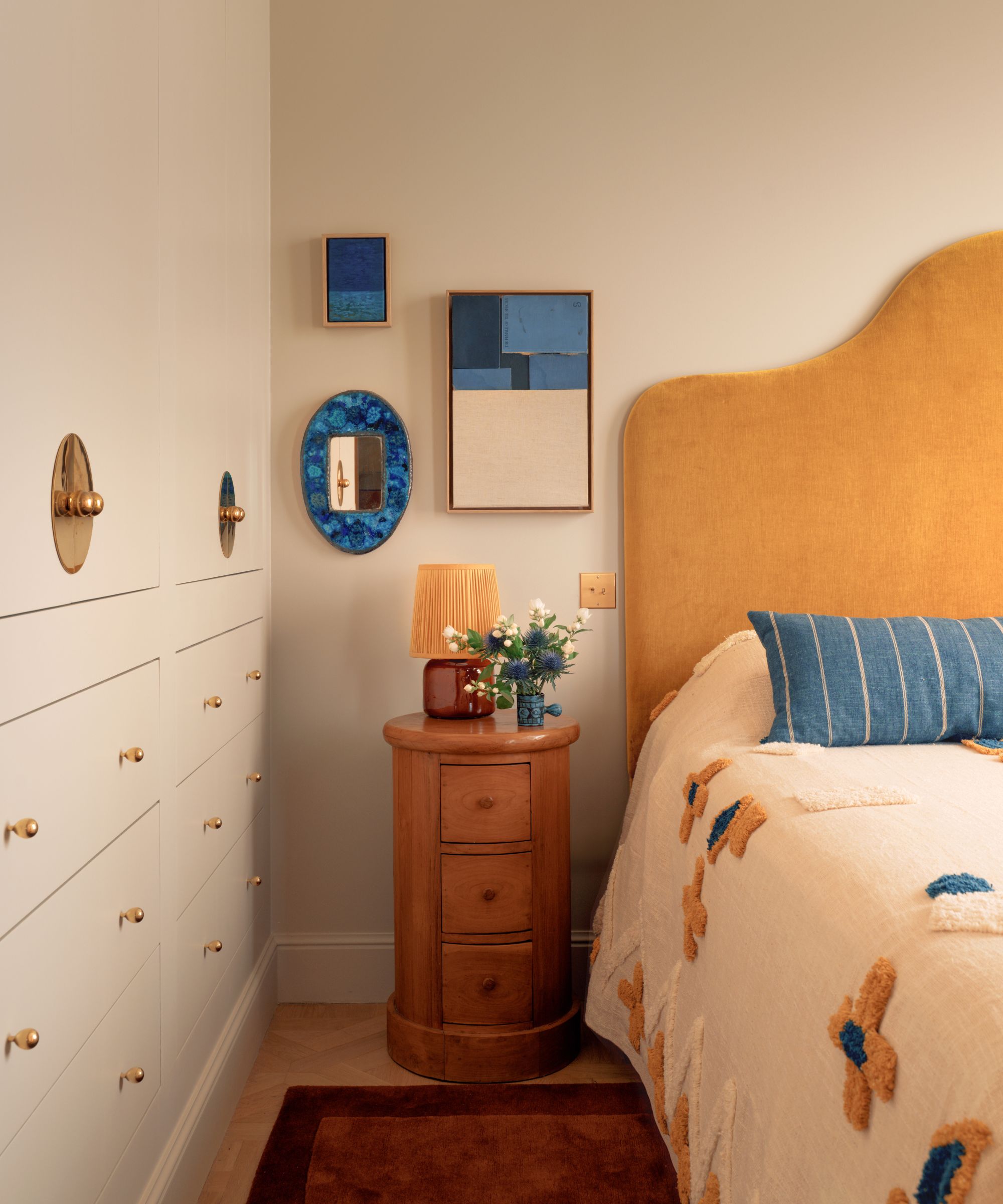 A bedroom with warm white walls, a mustard yellow headboard, a blue cushion, and artwork on the wall.