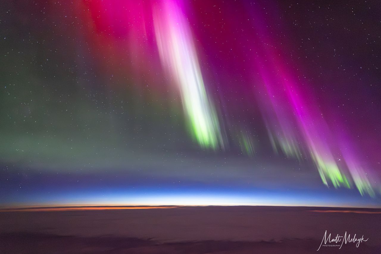 curtains of magenta and green light dance in the sky, a thin blue line marking Earth's atmosphere is visible in the distance.