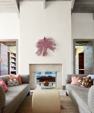 A light cream living room with two opposing sofas and a purple feather art piece hanging centrally above a fire place