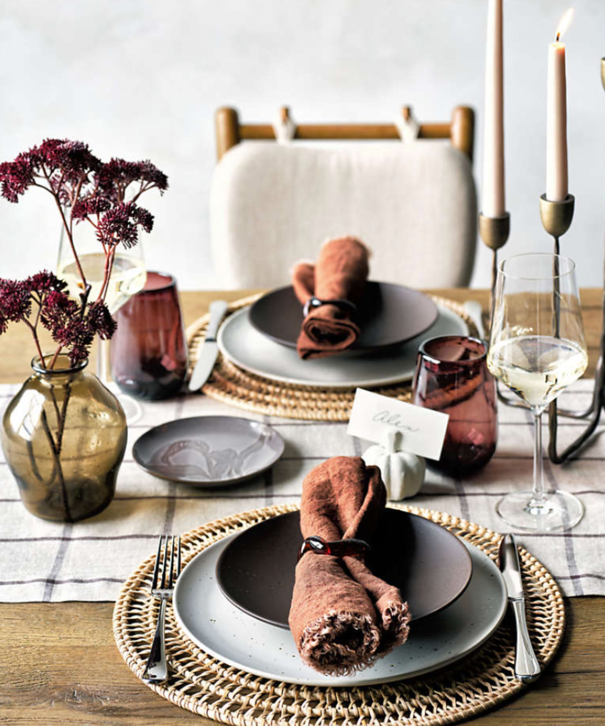 Table setting with woven placemats, gray plates, brown napkins and plum drinking glasses plus taper candles and dried flowers