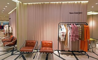 A clothing store with clothing hanging on rails, brown leather chairs and floor to ceiling pink curtains.