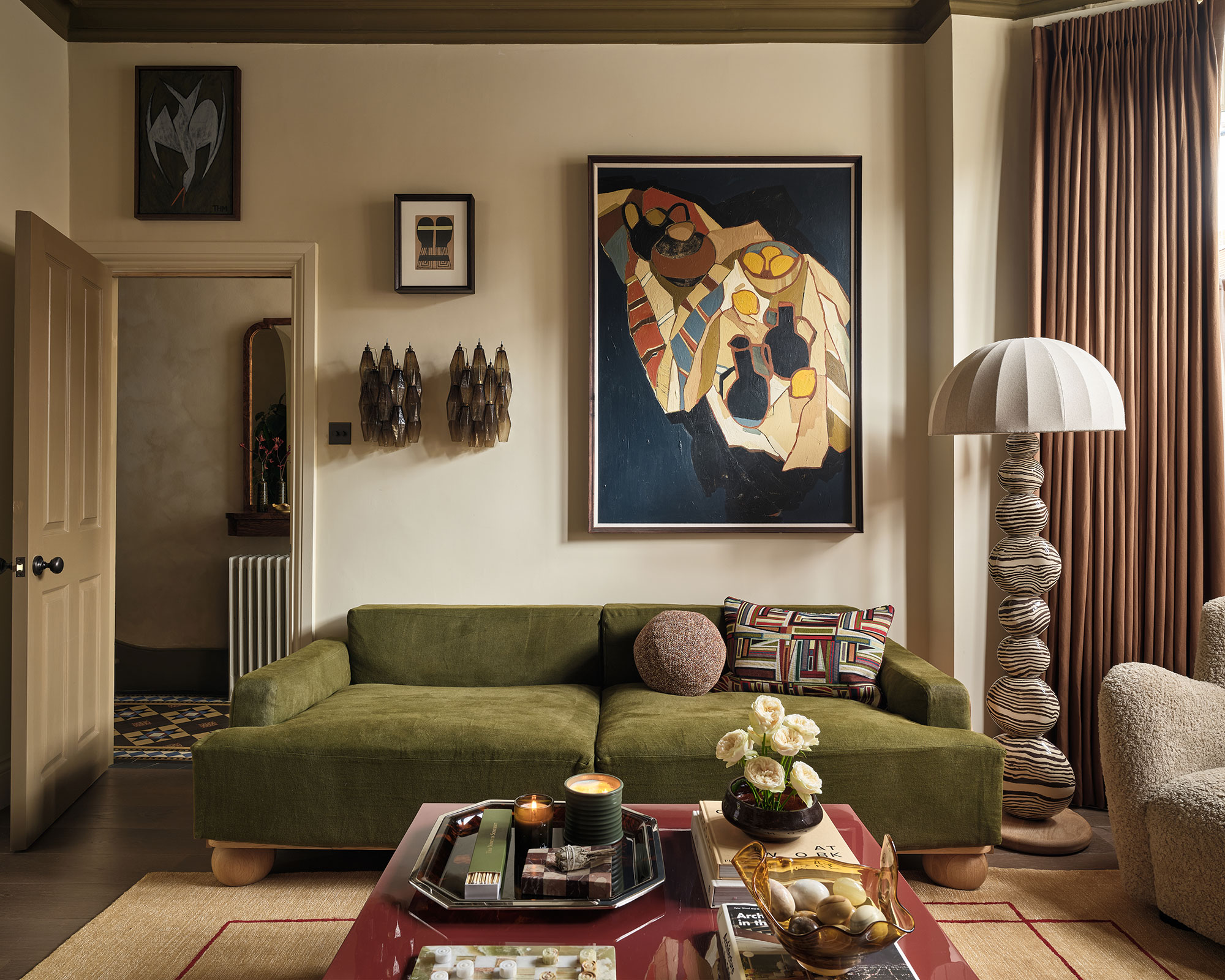 A warm, mid-century modern living room featuring an olive green velvet sofa, statement abstract art, an eclectic stacked-pebble floor lamp, and cozy earth tones with rich, textured decor.