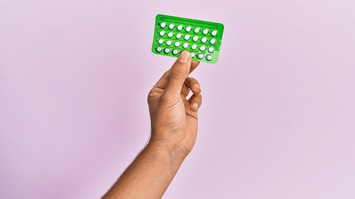 Young hispanic hand holding birth control pills over isolated pink background.