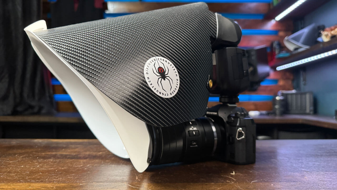 A camera with a carbon fiber-style diffuser attached, set on a wooden surface with a blurred background of tools and equipment