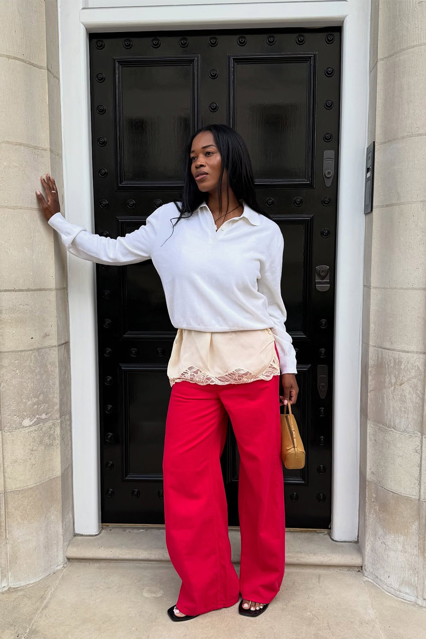 A sweater styling trend is shown in a photo of a woman&#039;s outfit where she&#039;s standing in a doorway outside wearing a white polo sweater over a cream lace slip dress, and red jeans worn with black sandals and a brown barrel bag
