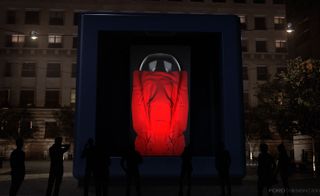 View of Ford's 'Favilla: To Every Light a Voice' installation - a large blue box with an object inside that is covered with red fabric. There is a building behind the box and silhouettes of people in front