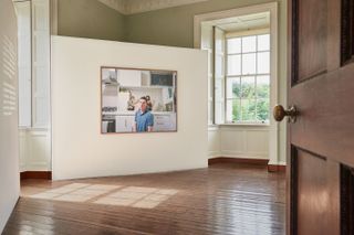 Inside an exhibition room with an image of a male wearing a blue polo shirt sat in a kitchen . Behind him is a landscape mirror with the reflection of three people posing.