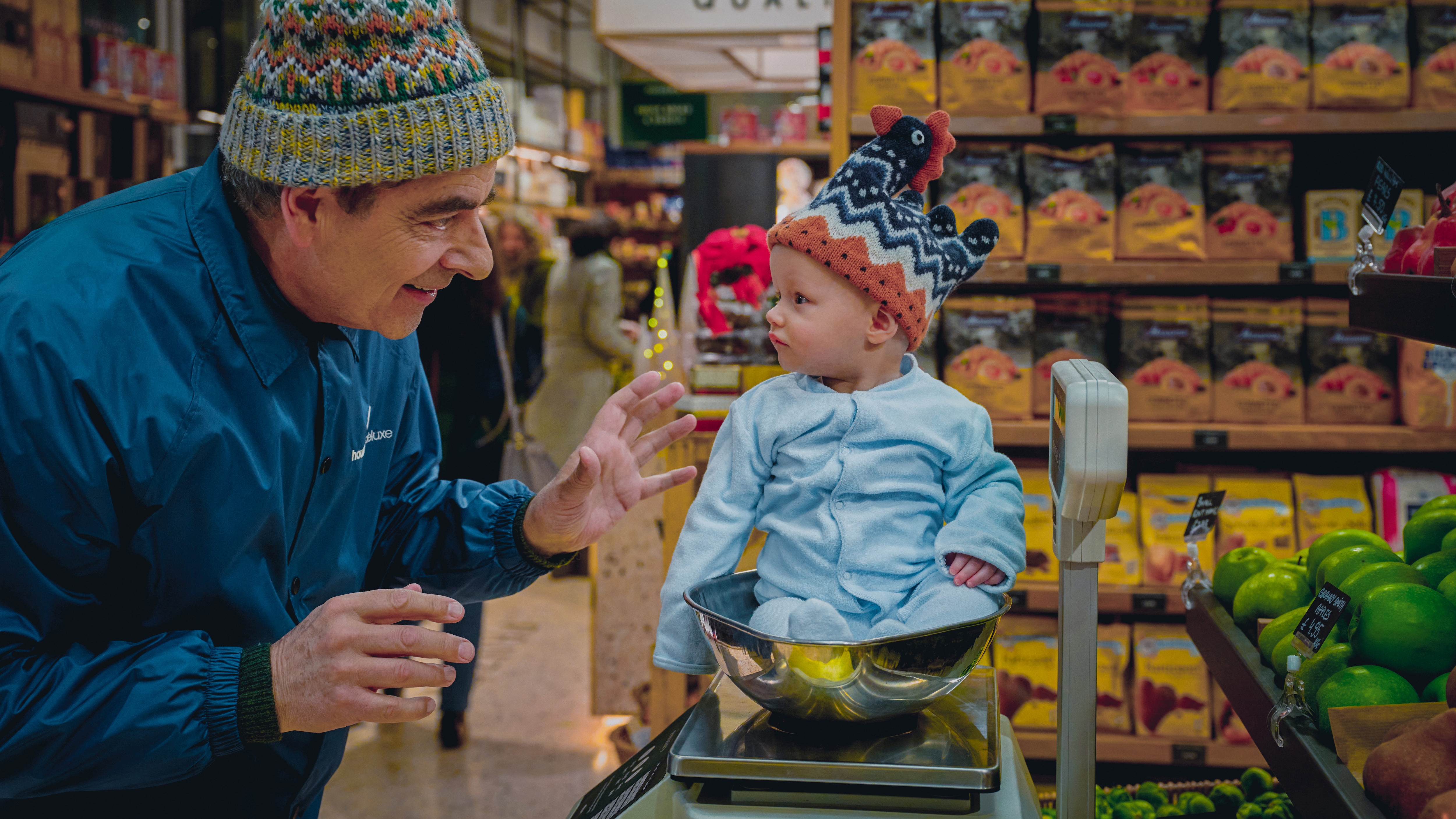 Rowan Atkinson as Trevor Bingley putting a baby in a winter hat and blue pajamas in a grocery store scale in the movie Man Vs Baby