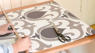 Person measuring patterned wallpaper on pasting table with other wallpaper tools