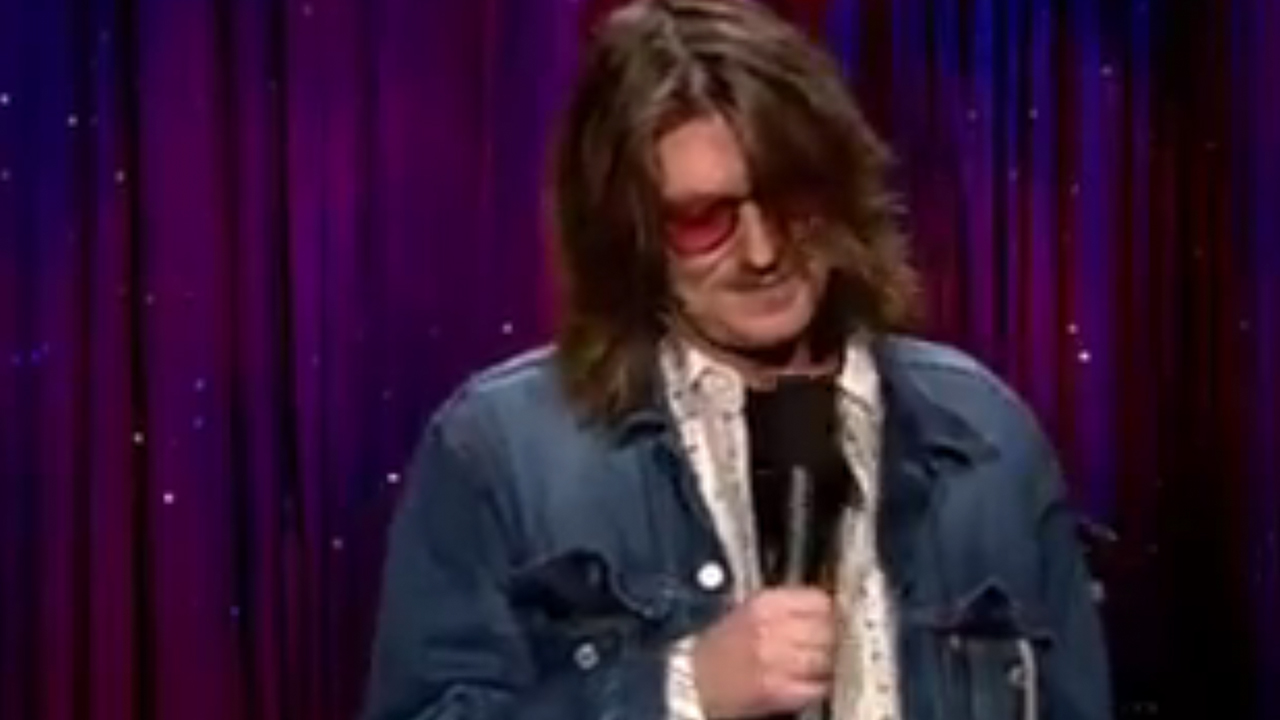 Mitch Hedberg wearing sunglasses and a jean jacket while performing on Late Night With Conan O'Brien.