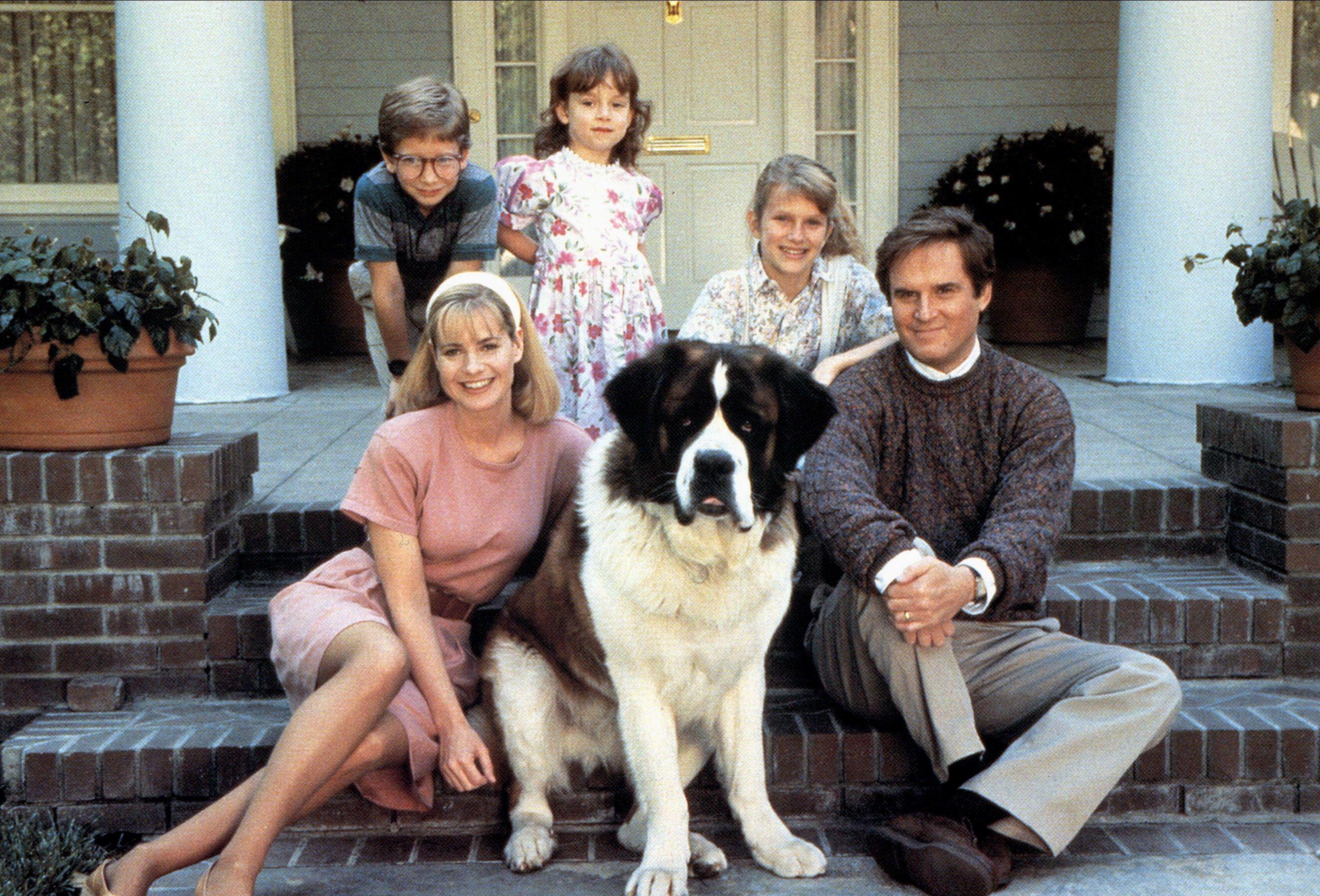 A large St Bernard dog sits on the front steps of a house with a smiling family gathered around it, including two adults and three children.