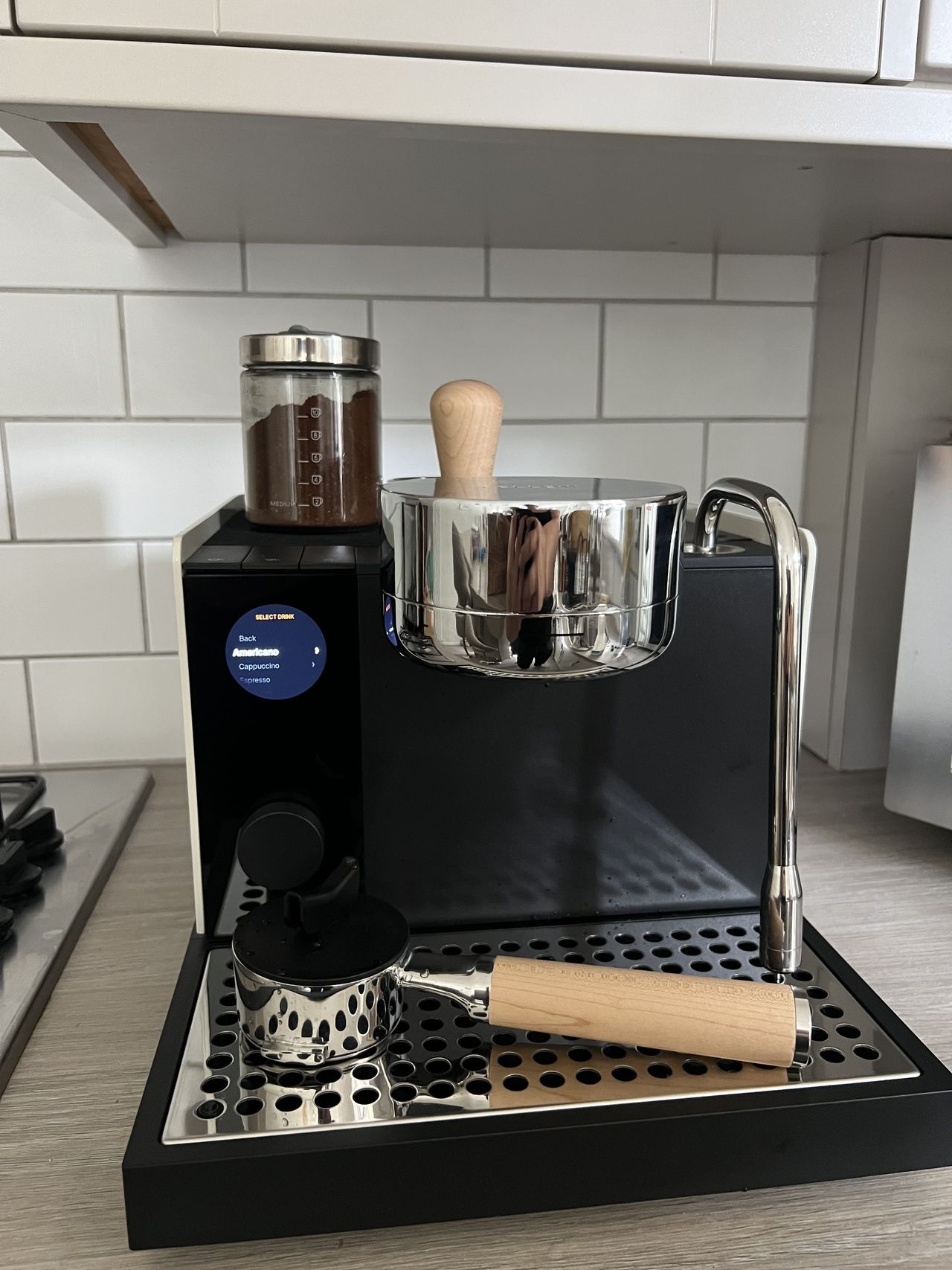 Image of the Fellow series 1 espresso machine on a light wood countertop in a white kitchen. There is a portafilter with a wooden handle on the base of the machine.