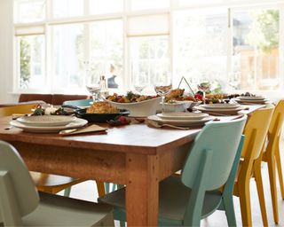 Wooden dining table set for meal, including food. Colourful chairs around it.