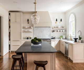 An L-shaped kitchen with a small kitchen island at the center. The cabinetry is a warm neutral pink color with white countertops, while the island is made from wooden cabinetry and a black countertop. The range cooker sits on the back wall with a plastered rangehood and two floating shelves either side.