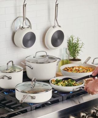 A set of KitchenAid cookware on a stove; hands sauteing vegetables and making ratatouille; against a white tiled wall.